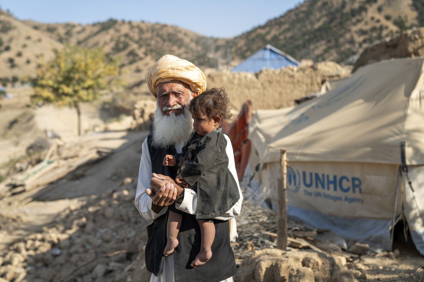 A man carrying a young child stands in front a UNHCR tent.